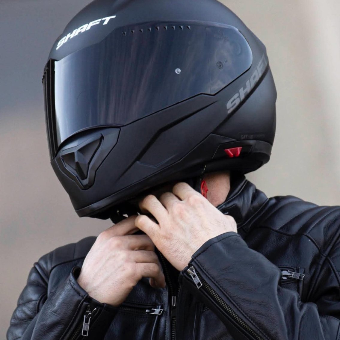 Motociclista ajustando la correa de seguridad de un casco integral negro Shaft con visera oscura, vistiendo chaqueta de cuero negra