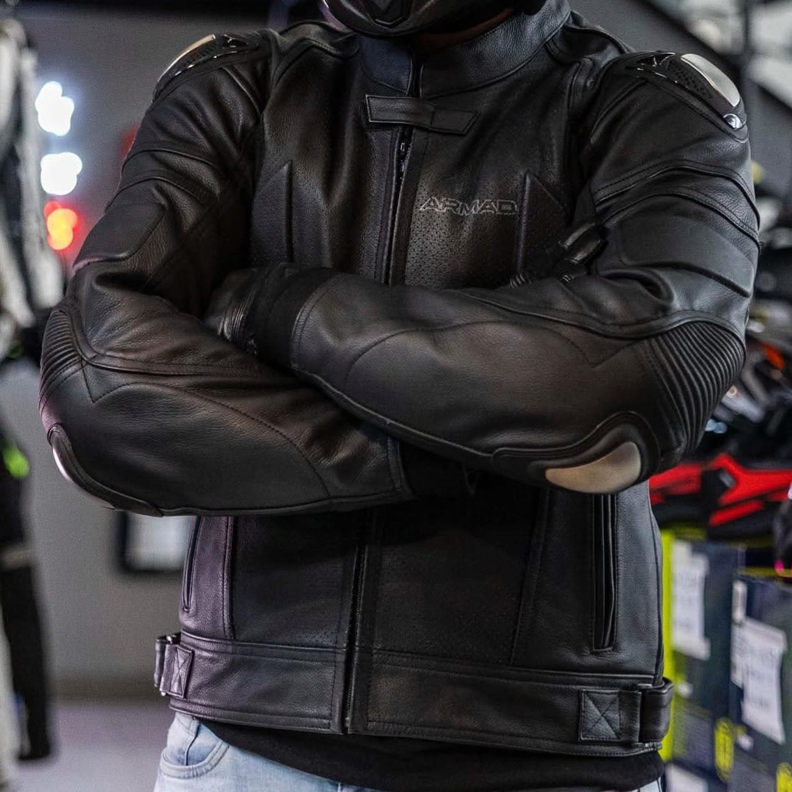 Chaqueta de moto de cuero negra ARMAG con protecciones integradas en codos y hombros, motorista con brazos cruzados en tienda de equipamiento